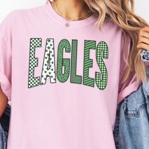 May include: A light pink short-sleeve t-shirt featuring the word EAGLES printed in large, block-style letters. Each letter has a unique green and white pattern, including checkered, star, leopard print, plaid, and diagonal stripe designs.