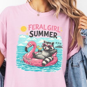 Könnte beinhalten: Hellrosa T-Shirt mit dem Text "FERAL GIRL SUMMER" und einer Grafik eines Waschbären mit Sonnenbrille, der sich auf einem rosa Flamingo-Schwimmring im Wasser entspannt. Der Waschbär hält einen Cocktail. Der Hintergrund zeigt einen sonnigen Himmel und Palmen.