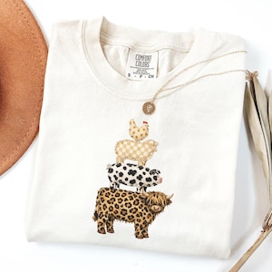 May include: Cream-colored Comfort Colors t-shirt featuring a stack of farm animals: a chicken, a sheep, a pig, and a Highland cow. A gold necklace with a pendant and a brown hat are also in the image.