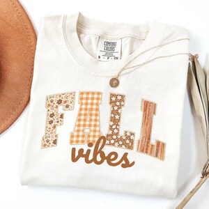 May include: A cream-colored short-sleeve t-shirt featuring the text FALL vibes in a mix of floral, orange gingham, and striped patterns. The shirt has a crew neckline and a Comfort Colors brand label at the inner collar, with a gold necklace resting on top.