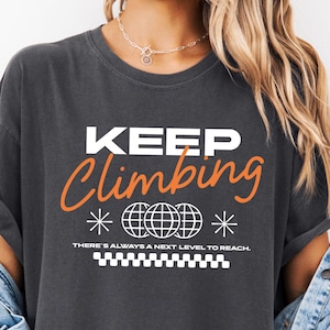 May include: Dark gray t-shirt with the words "KEEP Climbing" in white and orange script. The shirt features globe graphics and the text "THERE'S ALWAYS A NEXT LEVEL TO REACH." The shirt has rolled-up sleeves.