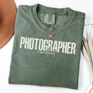 May include: An olive green t-shirt with the words "PHOTOGRAPHER assistant" printed in white. A gold necklace with a pendant rests on the shirt. A brown hat and dried plant are also in the image.