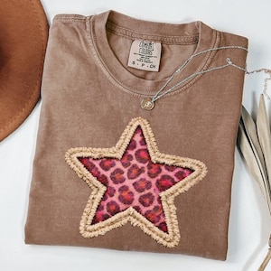 May include: Brown Comfort Colors t-shirt with a large star applique. The star features a pink leopard print center and a tan fringed border. A silver necklace with a small pendant rests on the shirt. The shirt is size S-CH.
