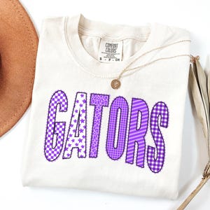 May include: A cream-coloured short-sleeve t-shirt with the word GATORS printed in purple. Each letter features a different pattern, including checkers, stars, leopard print, grid, and stripes. The shirt has a Comfort Colors brand label at the neck.