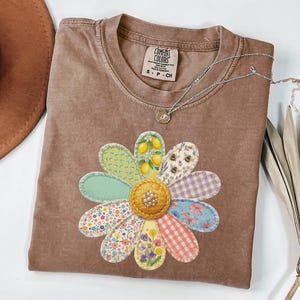 Op de afbeelding: Een bruin t-shirt met korte mouwen voorzien van een grote bloemafbeelding bestaande uit diverse stoffen bloemblaadjes met patronen, waaronder citroenen, bijen, ruitjes en bloemen, met een textuurrijk geel hart en een Comfort Colors label.