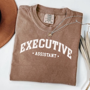 以下が含まれることがあります： 「EXECUTIVE ASSISTANT」の白いブロック文字がプリントされた茶色のTシャツ。Tシャツはクルーネックで、首元に小さなタグが付いています。ペンダント付きのシルバーのネックレスがTシャツの上にかけられています。