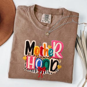 Puede incluir: Una camiseta marrón Comfort Colors con la palabra "Motherhood" en letras bordadas de colores. El diseño incluye un estampado de leopardo, corazones y un lazo rojo. Un collar plateado con un colgante descansa sobre la camiseta.