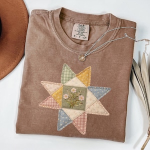 Może przedstawiać: Brązowa koszulka Comfort Colors z patchworkiem w kształcie gwiazdy. Patchwork zawiera różne pastelowe tkaniny i kwiatowy środek. Srebrny naszyjnik z małą zawieszką spoczywa na koszulce.
