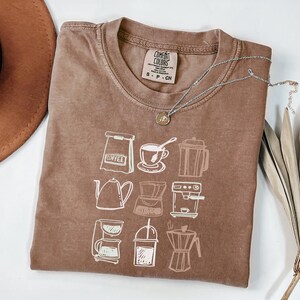 May include: A brown short-sleeved t-shirt featuring a white graphic print of various coffee brewing equipment, including a bag, cup, press, kettle, and espresso machine. The inner neck label reads Comfort Colors, S-P-CH, 100% Cotton, Made in Honduras.