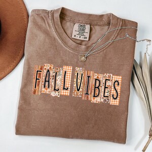 以下が含まれることがあります： 茶色のComfort Colors Tシャツ。装飾的なデザインで「FALL VIBES」の文字がプリントされています。オレンジ、茶色、クリーム色の花柄とチェック柄のデザイン。小さなペンダントが付いたシルバーのネックレスがシャツの上にかけられています。