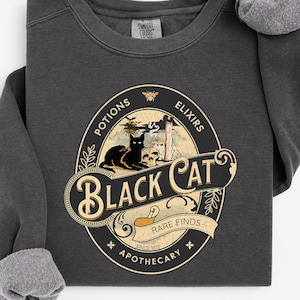 May include: A charcoal gray crewneck sweatshirt featuring a graphic print of a black cat, a skull, and apothecary-themed text that reads Black Cat Potions Elixirs Rare Finds Apothecary Since 1812, with a small bee icon at the top of the oval design.