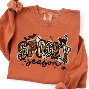 May include: A burnt orange long-sleeve shirt featuring the text SPOOKY season in black embroidery. The letters are filled with leopard, plaid, and checkered patterns, accented by embroidered bats, ghosts, a black cat, and a spider.
