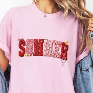 Pode incluir: Uma t-shirt de manga curta rosa claro com a palavra SUMMER em letras grandes, vermelhas e cor-de-rosa com padrões, decorada com pequenos gráficos de morangos e flores, usada sob uma camisa de ganga azul de manga comprida.