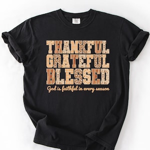 May include: A black short-sleeved t-shirt featuring the words THANKFUL GRATEFUL BLESSED in a tan and brown patterned font, with the text God is faithful in every season printed in a smaller, simple tan font directly underneath the main words.