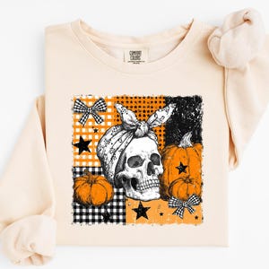 May include: A light beige crewneck sweatshirt featuring a graphic print of a skull wearing a patterned headscarf, surrounded by orange pumpkins, black stars, and black and white checkered bows on a background of orange and black squares.