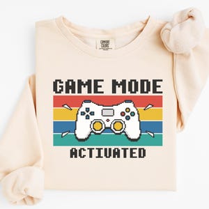 Puede incluir: Sudadera color crema con un diseño pixelado de mando de videojuegos. El diseño incluye el texto "GAME MODE ACTIVATED" encima y debajo del mando, sobre un fondo de rayas horizontales rojas, amarillas, azules y turquesas.