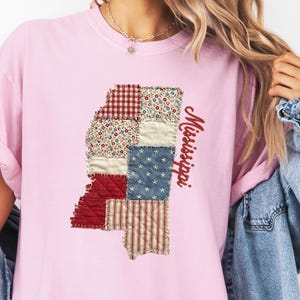 May include: A light pink t-shirt featuring a patchwork design of the state of Mississippi. The patchwork includes red gingham, floral, star-spangled, and striped patterns. The word "Mississippi" is printed in red script.