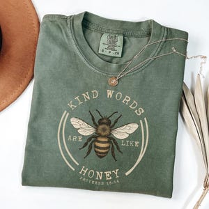Op de afbeelding: Een olijfgroen t-shirt met de woorden "Kind Words Are Like Honey" rond een gedetailleerde illustratie van een bij. Het shirt heeft ook de tekst "Proverbs 16:24". Een zilveren ketting met een kleine hanger en een bruine hoed zijn ook zichtbaar.