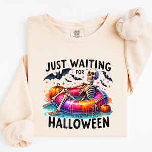 May include: A light beige crewneck sweatshirt featuring a graphic of a skeleton lounging in a colorful, melting-effect pool float. The design includes orange pumpkins, black bats, and the text "JUST WAITING FOR HALLOWEEN" in black lettering.