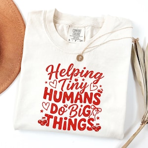 Peut inclure: T-shirt crème à col rond avec le texte "Helping Tiny Humans Do Big Things" en rouge. Le motif comprend des cœurs, des étoiles et des cerises. Un collier doré avec un pendentif et un chapeau marron sont également visibles.