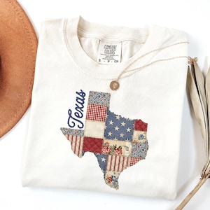 Könnte beinhalten: Cremefarbenes Comfort Colors Sweatshirt mit einem Texas-Karten-Design im Patchwork-Stil. Die Karte enthält rote, weiße und blaue Muster, einschließlich Sterne und Streifen. Das Wort "Texas" ist in Marineblau geschrieben. Eine goldene Halskette und ein brauner Hut sind ebenfalls zu sehen.