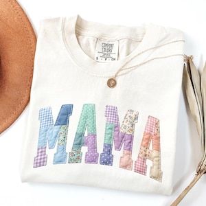 Puede incluir: Sudadera color crema con la palabra "MAMA" aplicada en tela de patchwork. Las letras de patchwork presentan varios patrones y colores, incluyendo cuadros azules, estampados florales y lunares lavanda. También se ven un collar dorado y un sombrero marrón.