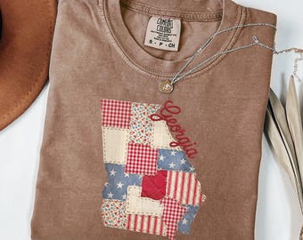 Może przedstawiać: Brązowa koszulka Comfort Colors z patchworkowym wzorem stanu Georgia. Patchwork zawiera czerwone, białe i niebieskie wzory. Widoczny jest również srebrny naszyjnik i brązowy kapelusz.