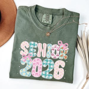 May include: An olive green Comfort Colors t-shirt with the text "Senior 2026" in a patchwork design with pink bows and floral accents. A silver necklace and a brown hat are also visible.