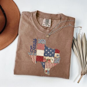 May include: A brown t-shirt featuring a patchwork design of the state of Texas. The Texas outline is filled with various patterns, including stars, stripes, and floral prints. The word "Texas" is embroidered in blue script. A silver necklace and a brown hat are also visible.