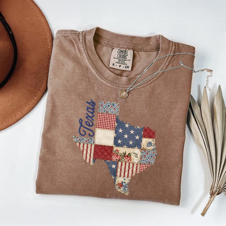 May include: A brown t-shirt featuring a patchwork design of the state of Texas. The Texas outline is filled with various patterns, including stars, stripes, and floral prints. The word "Texas" is embroidered in blue script. A silver necklace and a brown hat are also visible.