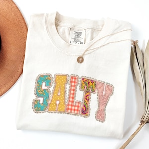 May include: A cream-colored short-sleeve t-shirt featuring the word SALTY in large, block-style letters. Each letter has a unique pattern: floral, yellow polka dot, orange gingham, paisley, and pink damask, all outlined in a silver glitter border.