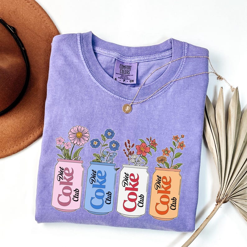 May include: A lavender Comfort Colors t-shirt featuring four Diet Coke cans, each filled with different flowers. The cans are pink, blue, white, and orange. A gold necklace and a brown hat are also visible.