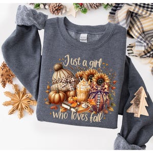 May include: Dark gray sweatshirt with a fall-themed graphic. The graphic includes a knitted hat, pumpkins, sunflowers, a football, and a beverage. Text on the sweatshirt reads "Just a girl who loves fall."
