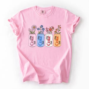 May include: A light pink t-shirt featuring a graphic of four Diet Coke cans, each holding a different arrangement of flowers. The cans are in pink, blue, white, and orange, with the words "Diet Coke Club" printed on them.