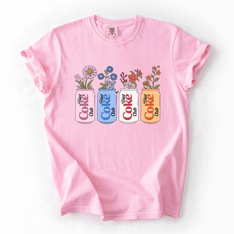 May include: A light pink t-shirt featuring a graphic of four Diet Coke cans, each holding a different arrangement of flowers. The cans are in pink, blue, white, and orange, with the words "Diet Coke Club" printed on them.
