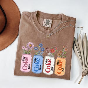 May include: Brown t-shirt with a floral design. The graphic features four Diet Coke cans in pink, blue, white, and orange, each with a different type of flower. A silver necklace and a brown hat are also visible.