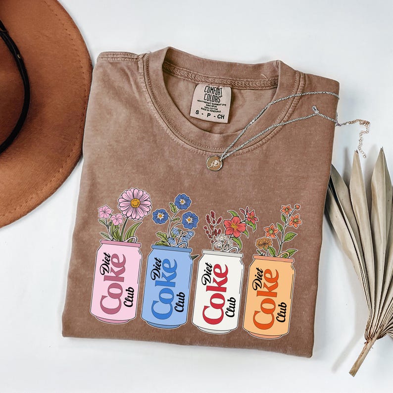 May include: Brown t-shirt with a floral design. The graphic features four Diet Coke cans in pink, blue, white, and orange, each with a different type of flower. A silver necklace and a brown hat are also visible.
