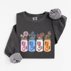 May include: Dark gray sweatshirt featuring four Diet Coke cans with floral arrangements. The cans are pink, blue, white, and orange, each with a different flower type. The text "Diet Coke Club" is on each can.