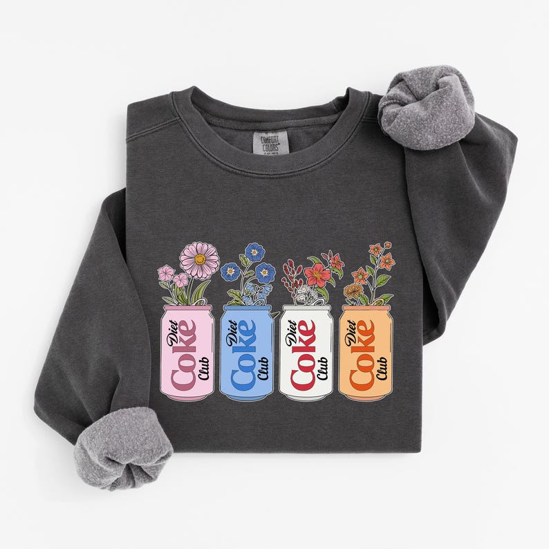 May include: Dark gray sweatshirt featuring four Diet Coke cans with floral arrangements. The cans are pink, blue, white, and orange, each with a different flower type. The text "Diet Coke Club" is on each can.