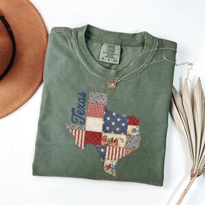 May include: An olive green t-shirt featuring a patchwork design of the state of Texas. The Texas outline is filled with various patterns, including stars, stripes, and floral prints. The word "Texas" is embroidered in blue script. A silver necklace with a pendant rests on the shirt.