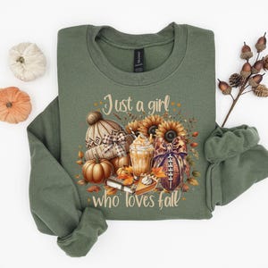 May include: Olive green sweatshirt with the text "Just a girl who loves fall." The graphic includes a knit hat, pumpkins, sunflowers, a football, and a pumpkin spice latte. The sweatshirt is folded.
