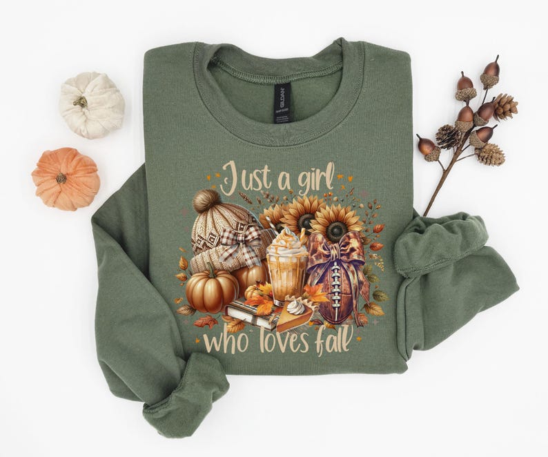 May include: Olive green sweatshirt with the text "Just a girl who loves fall." The graphic includes a knit hat, pumpkins, sunflowers, a football, and a pumpkin spice latte. The sweatshirt is folded.