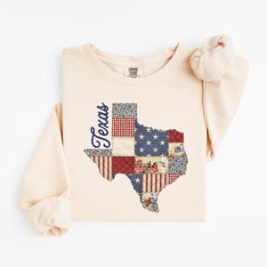 May include: Cream-colored long-sleeve sweatshirt featuring a patchwork design of the state of Texas. The Texas outline is filled with various patterns, including stars, stripes, and floral prints. The word "Texas" is written in a cursive font on the side.