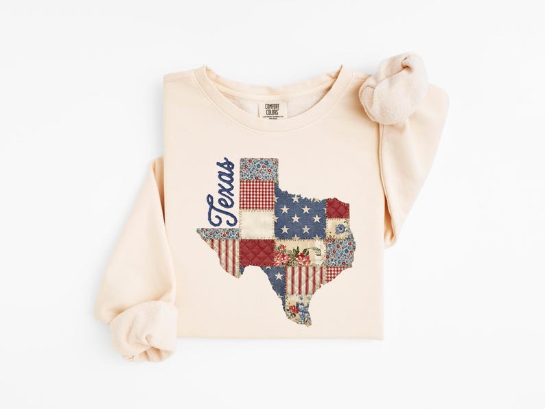 May include: Cream-colored long-sleeve sweatshirt featuring a patchwork design of the state of Texas. The Texas outline is filled with various patterns, including stars, stripes, and floral prints. The word "Texas" is written in a cursive font on the side.