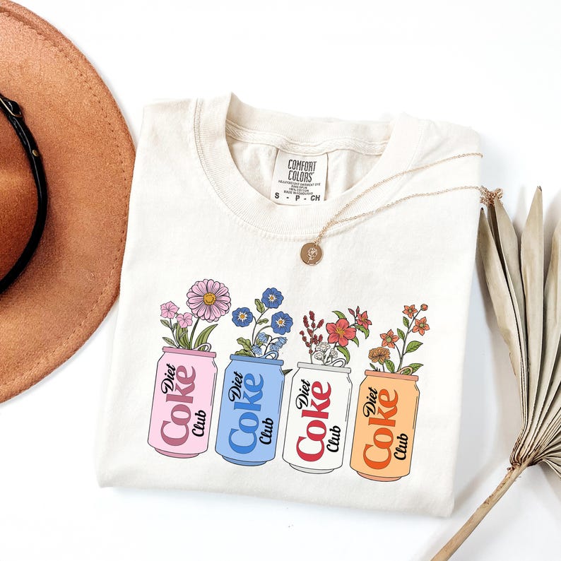 May include: Cream-colored sweatshirt featuring a graphic of four Diet Coke cans, each filled with different flowers. The cans are pink, blue, white, and orange, with the words "Diet Coke Club" printed on them. A gold necklace and a brown hat are also visible.
