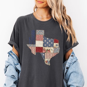 May include: Dark gray t-shirt featuring a patchwork design of the state of Texas. The Texas outline is filled with red, white, and blue patterns, including stars, stripes, and floral prints. The word "Texas" is written vertically in blue.