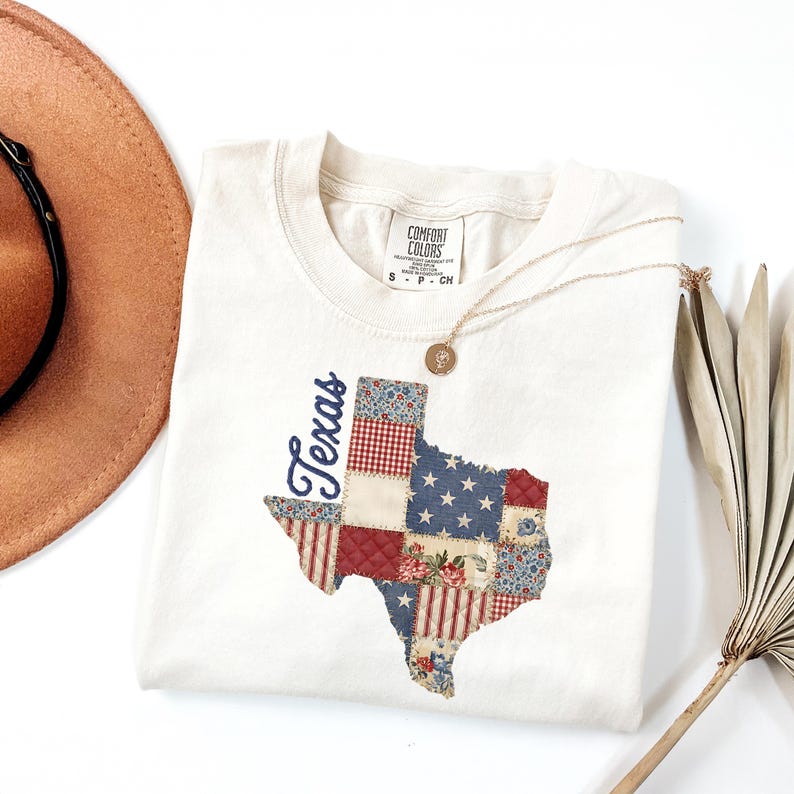 May include: Cream-colored Comfort Colors sweatshirt featuring a patchwork Texas map design. The map incorporates red, white, and blue patterns, including stars and stripes. The word "Texas" is written in a navy blue script. A gold necklace and a brown hat are also visible.