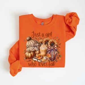 May include: Orange sweatshirt with a fall-themed graphic. The graphic includes a knit hat, pumpkins, sunflowers, a football, and a coffee drink. Text on the shirt reads "Just a girl who loves fall."