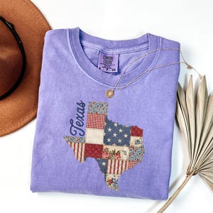 May include: A light purple Comfort Colors t-shirt featuring a patchwork design of the state of Texas. The Texas shape is made of various fabric patterns, including stars and stripes, floral, and gingham. A gold necklace and a brown hat are also in the image.