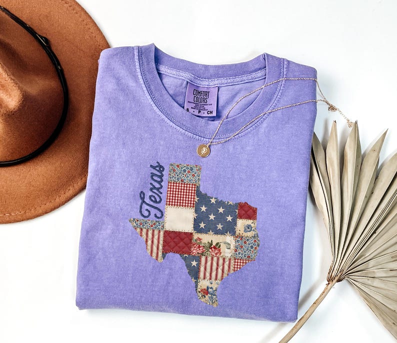 May include: A light purple Comfort Colors t-shirt featuring a patchwork design of the state of Texas. The Texas shape is made of various fabric patterns, including stars and stripes, floral, and gingham. A gold necklace and a brown hat are also in the image.